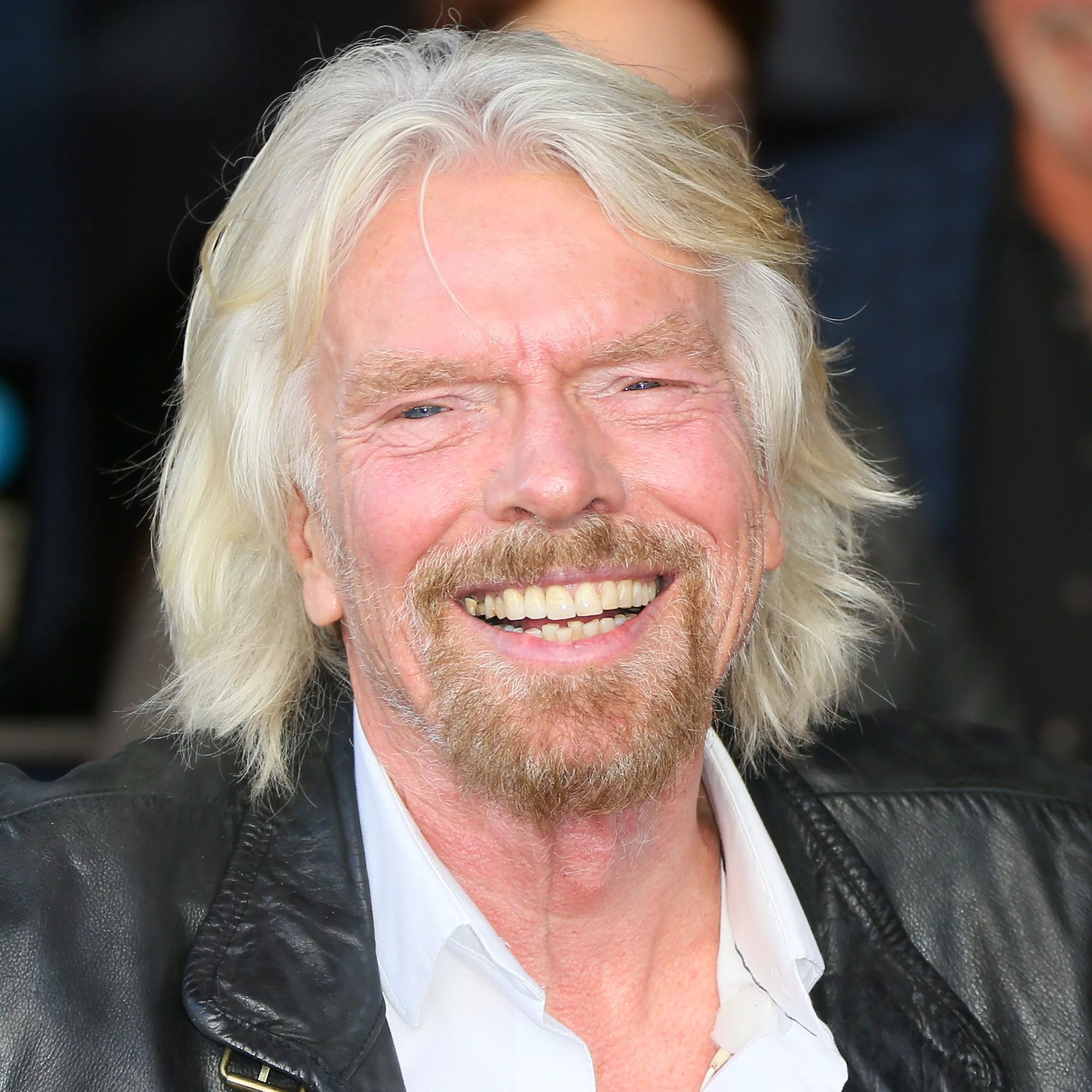 A smiling older man with long blond hair and a beard, wearing a leather jacket and a white shirt, exuding confidence and charisma in a professional setting.
